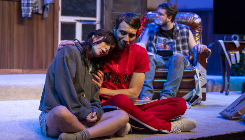 Two student actors sitting corss-legged on stage holding each other.