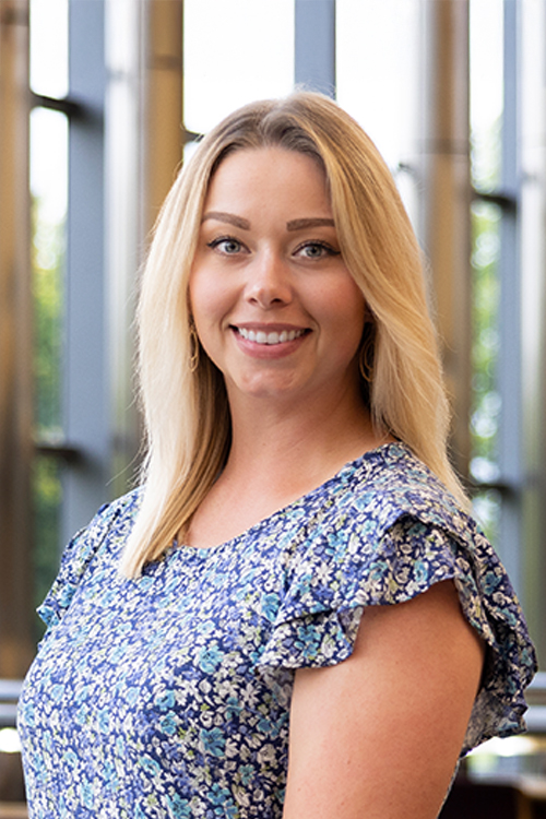 Kayla Bueby wearing a blue floral blouse, stands in a well-lit indoor setting.