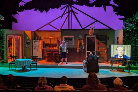 A darkened theatre stage with a set of a living room, Jimmy Carter playing on a television