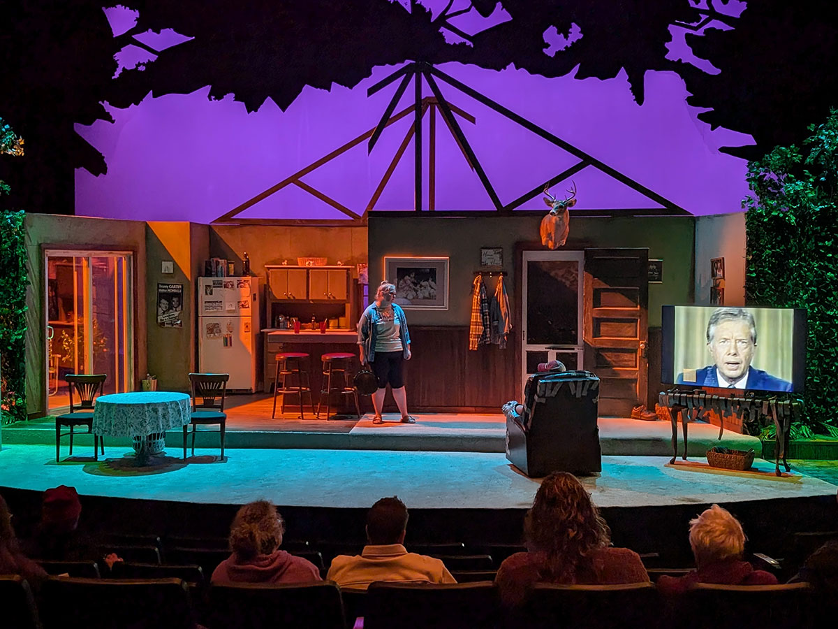 A darkened theatre stage with a set of a living room, Jimmy Carter playing on a television