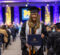 A graduate smiles and holds a diploma during a UM-Flint graduation ceremony in a large hall.