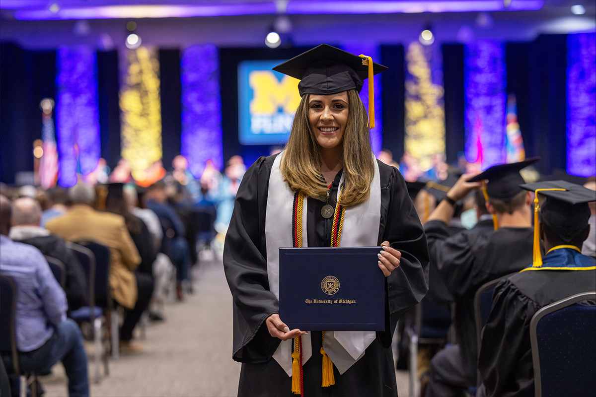 A graduate smiles and holds a diploma during a UM-Flint graduation ceremony in a large hall.