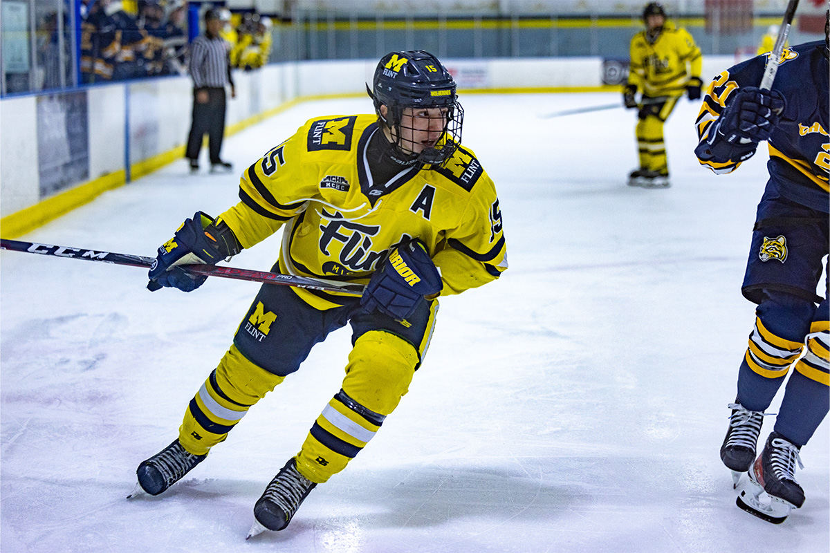 A hockey player in a yellow and navy uniform, marked with "Flint," skates on an ice rink during a game, gripping his stick.