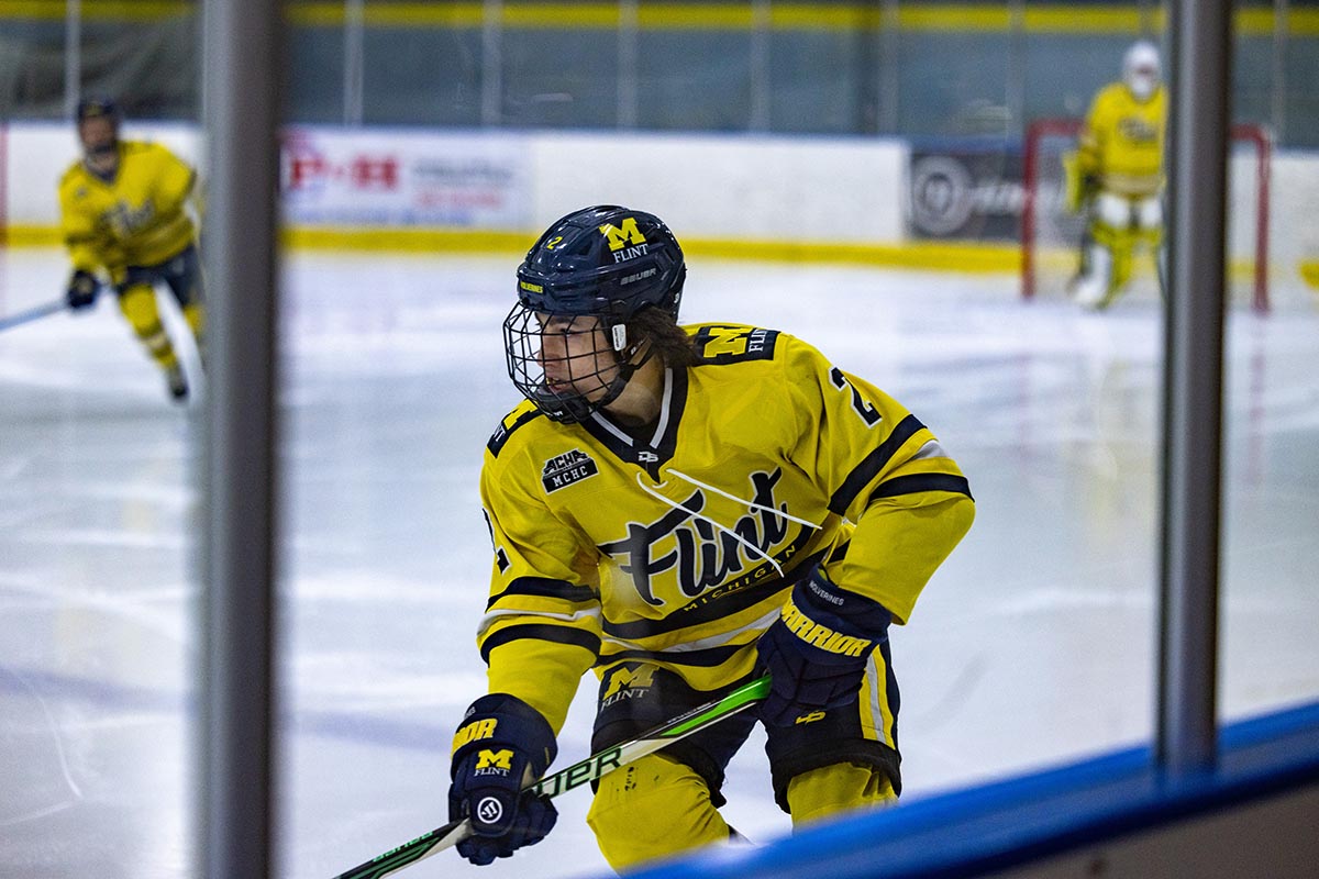 A UM-Flint hockey player on the ice photographed through the glass