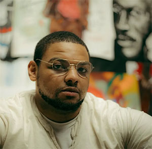 A young Black male artist posting in a cream colored t-shirt in front of a background featuring his artwork