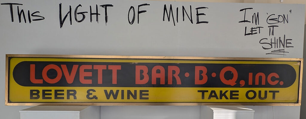 An installation in the exhibition that features a sign for Lovett Bar-B-Q with the phrase "This light of mine I'm gon let it shine" written on the wall above.