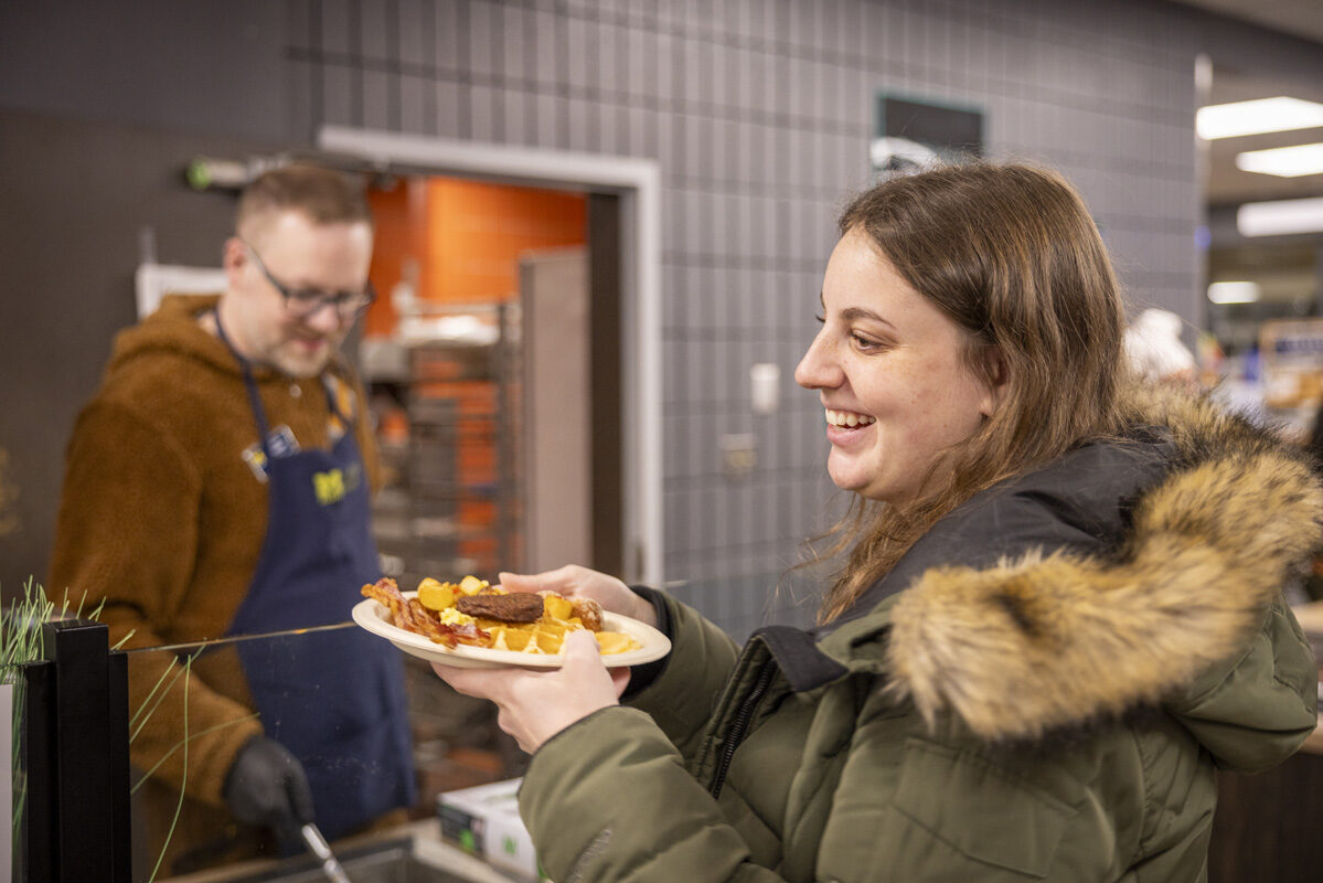 A smiling person holding a plate of breakfast food in a cafeteria setting, interacting with a staff member behind a counter.