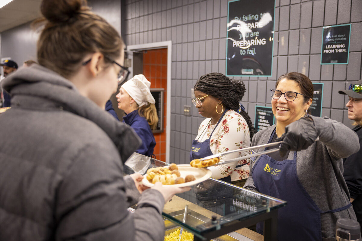 A cafeteria worker smiles while serving breakfast food to a customer.