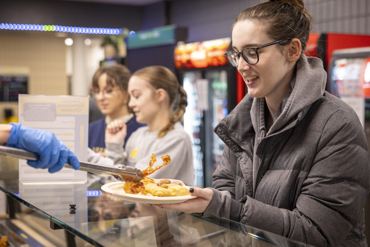 A smiling person holds a plate as a cafeteria worker serves their breakfast.