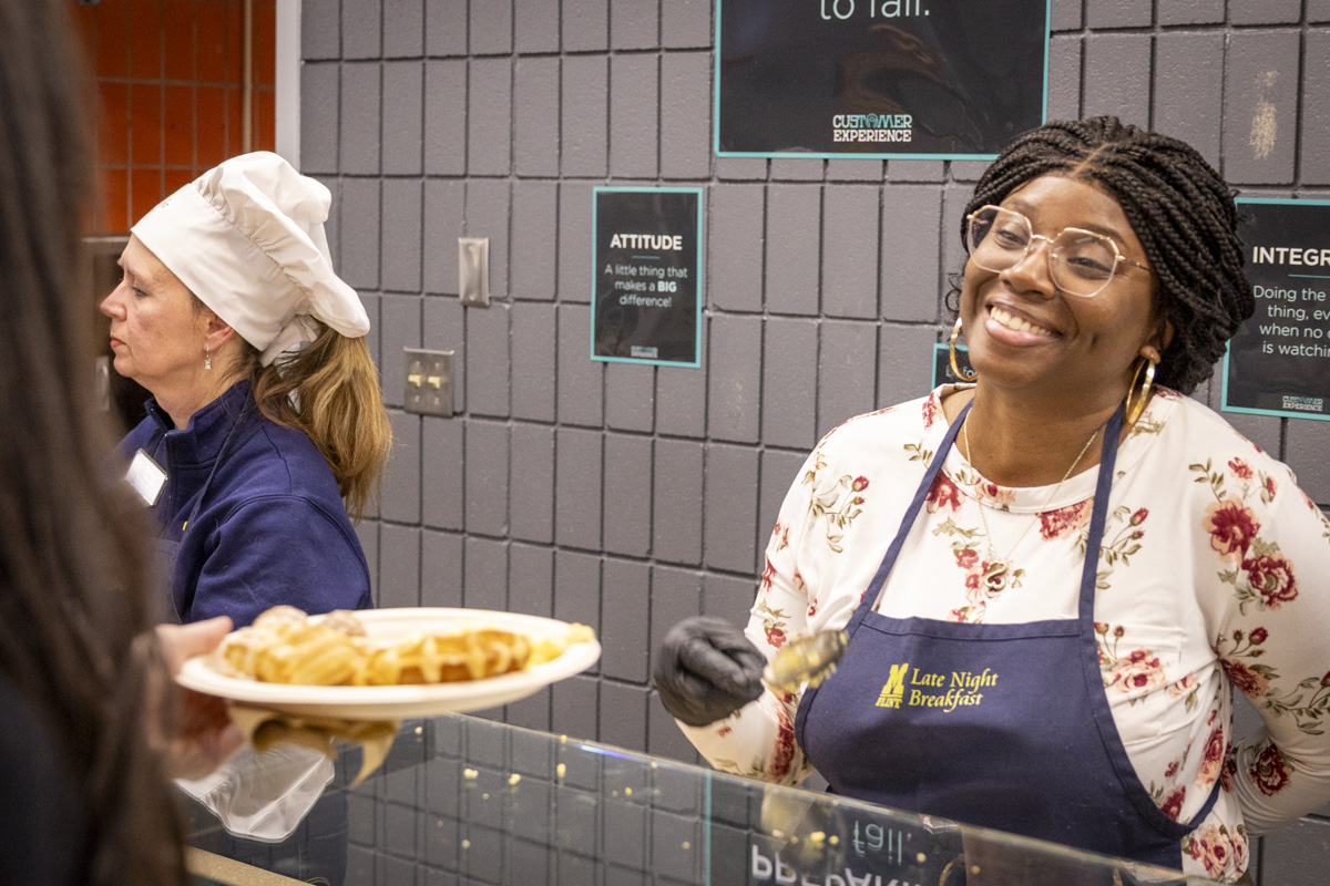 A smiling cafeteria worker serves food while another stands in the background.