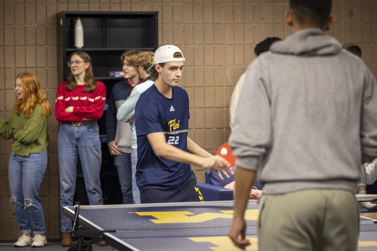 Two young people play ping pong while a group of peers watch in a casual setting.