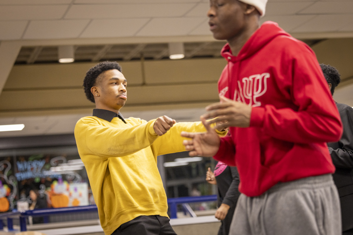 A young person in a yellow sweater gestures dynamically while another in a red hoodie stands nearby, both appearing to be mid-dance in a casual indoor setting.