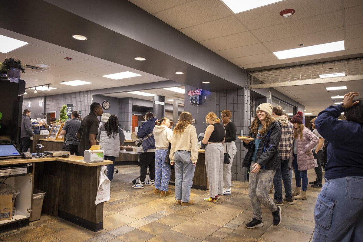 A busy cafeteria with people standing in line for breakfast, while one person smiles and walks with a tray of food.