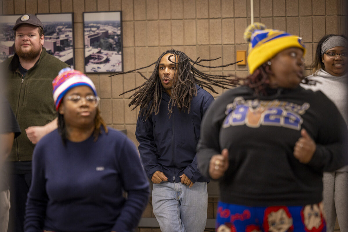 A group of people participate in an activity, including a person with long dreadlocks in motion and others wearing casual clothes and colorful hats.