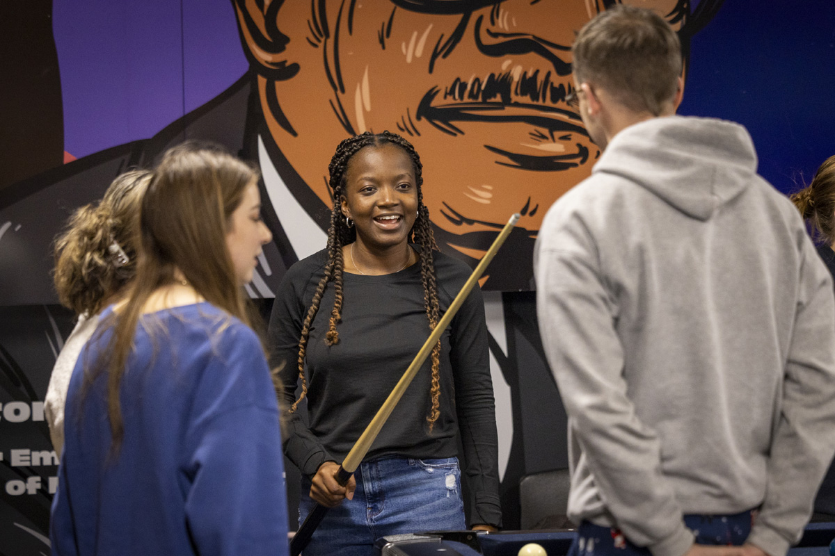 A young person holding a pool cue smiles while chatting with a group of people in front of a colorful mural.