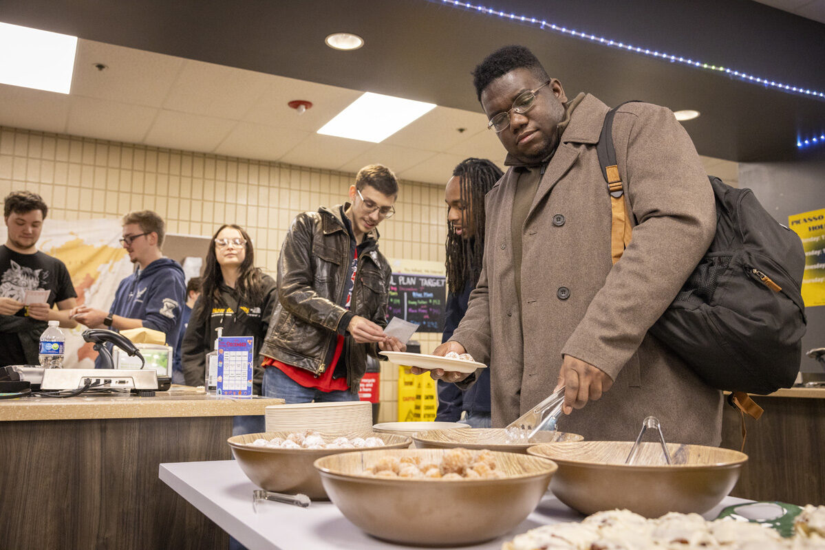 A person in a brown coat serves themself from a table of breakfast pastries while others wait in line behind them.