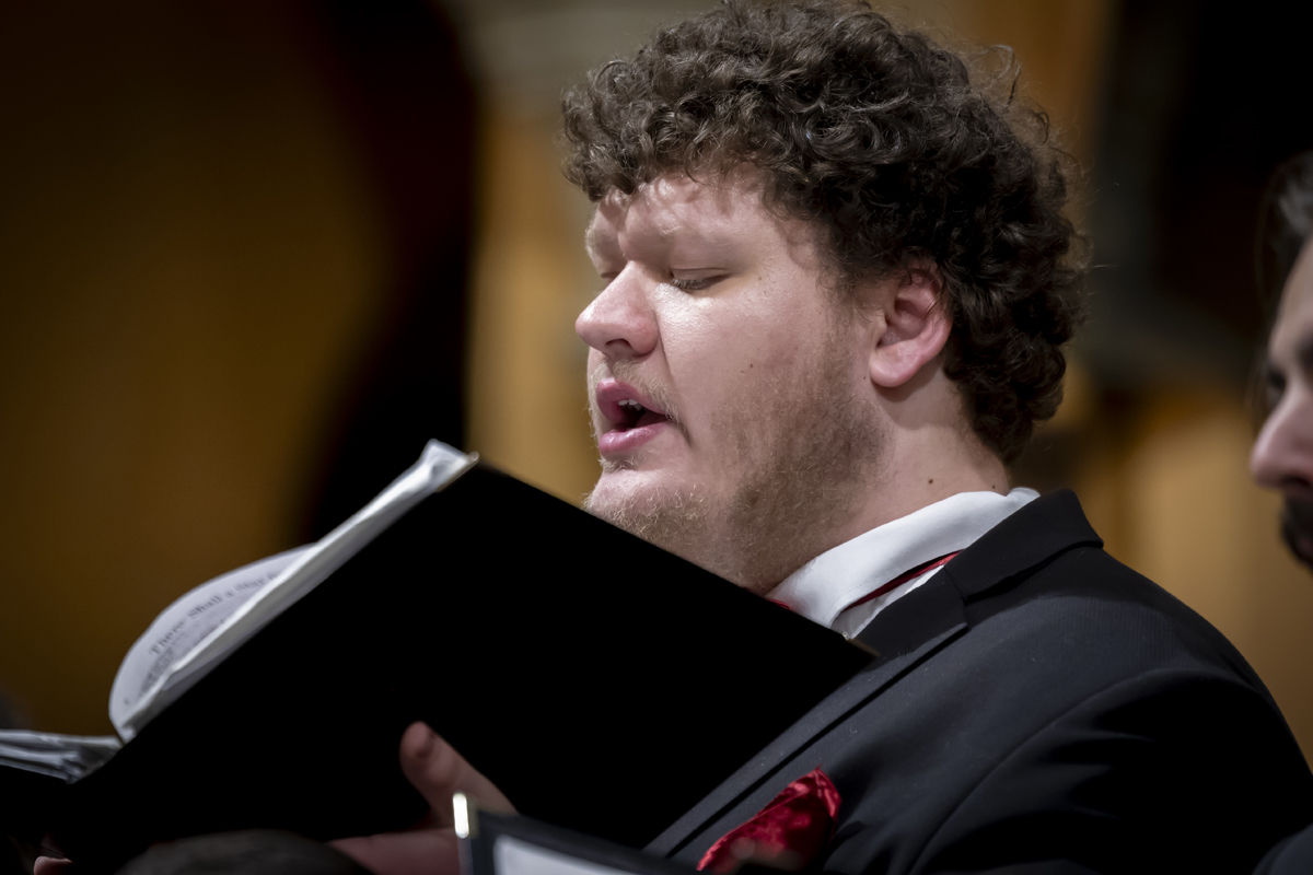 A man with curly hair sings while holding sheet music.