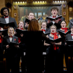 A choir dressed in black suits and dresses with red accents performs under the direction of a conductor in a wood-paneled church setting.