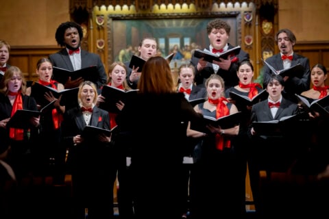 A choir dressed in black suits and dresses with red accents performs under the direction of a conductor in a wood-paneled church setting.