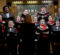 A choir dressed in black suits and dresses with red accents performs under the direction of a conductor in a wood-paneled church setting.