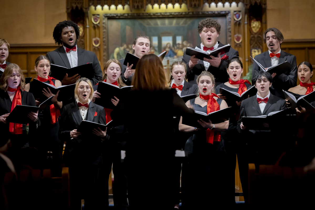 A choir dressed in black suits and dresses with red accents performs under the direction of a conductor in a wood-paneled church setting.