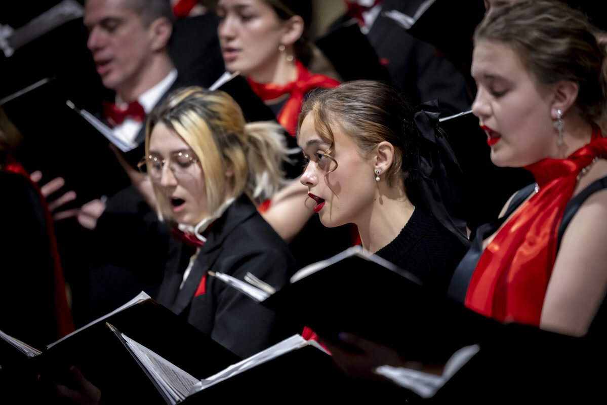 Choir members performing in formal attire with red accents, holding songbooks.