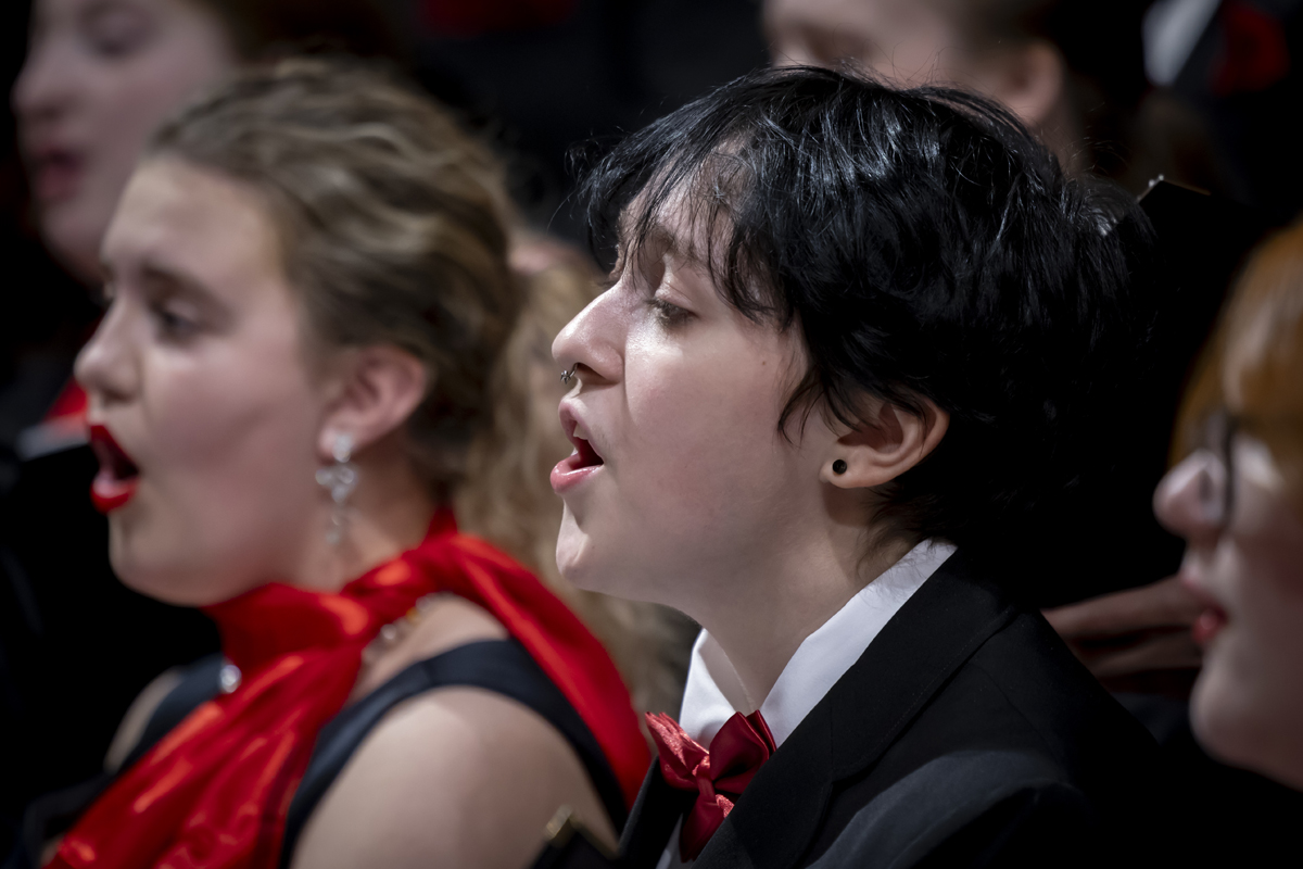 A diverse choir sings passionately, dressed in black with red accents.