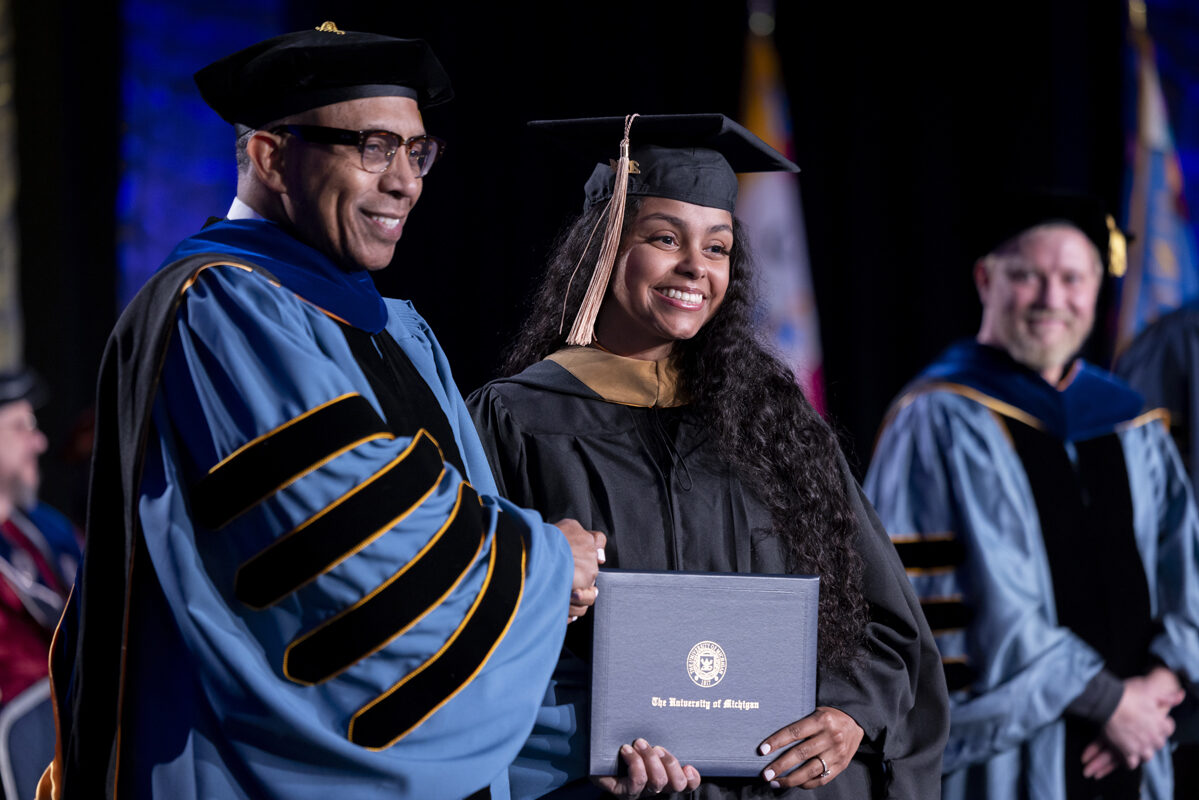 A smiling graduate shakes hands with a faculty member while holding her diploma.