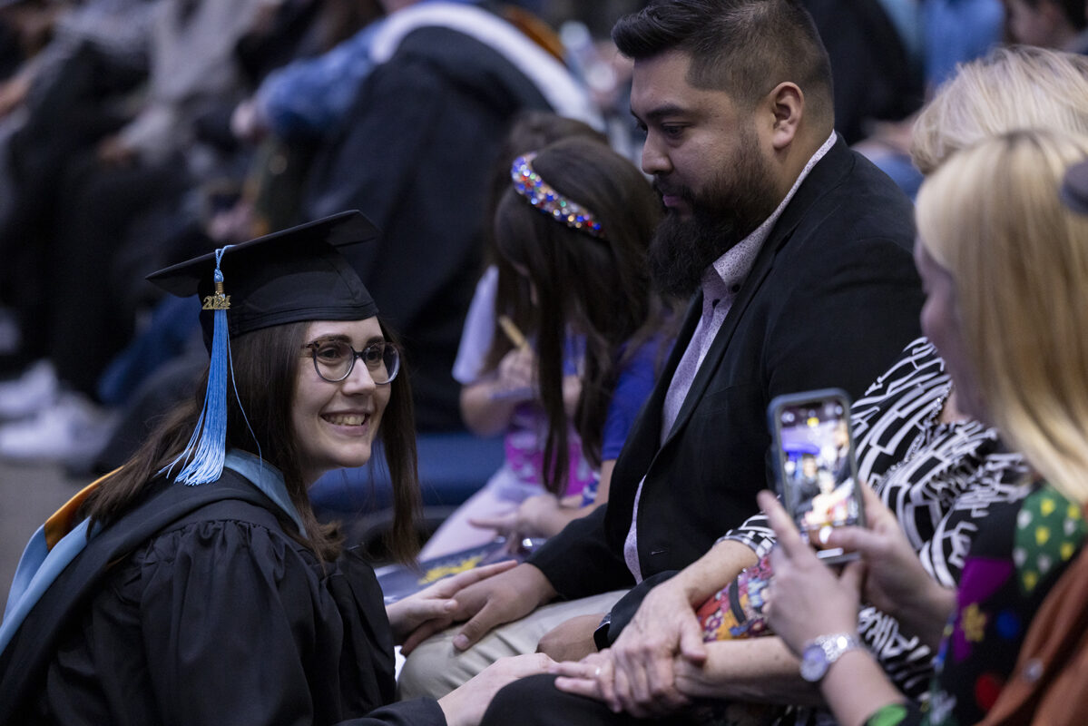 A smiling graduate in cap and gown kneels to hold hands with family members.