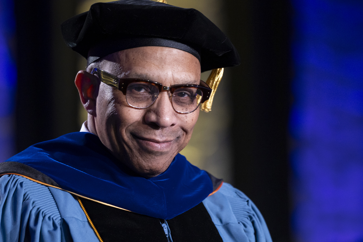 A smiling faculty member in academic regalia with glasses and a black cap.