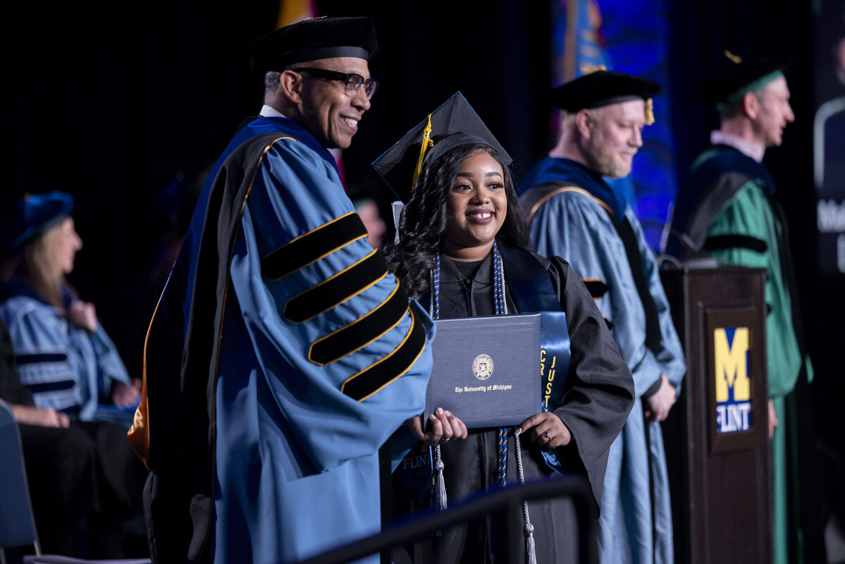 A smiling graduate holds her diploma while shaking hands with a faculty member.