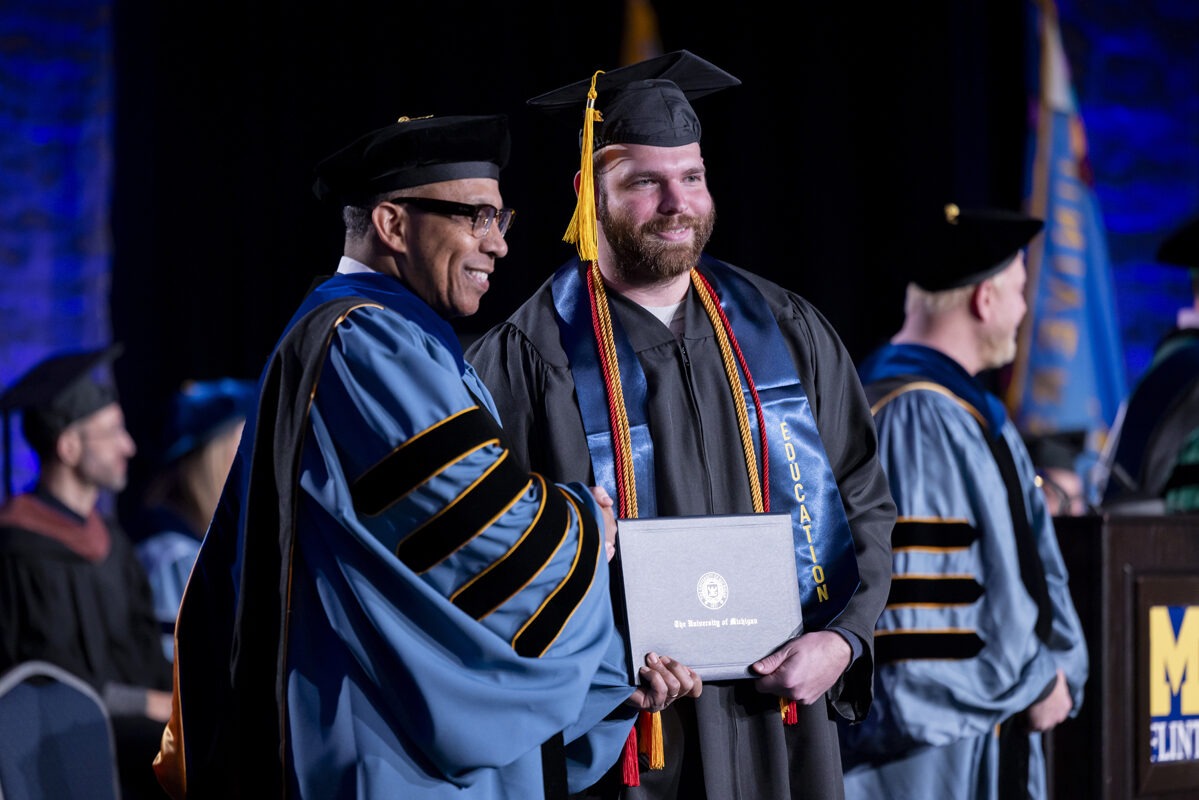 A graduate shakes hands with a faculty member while posing with his diploma.