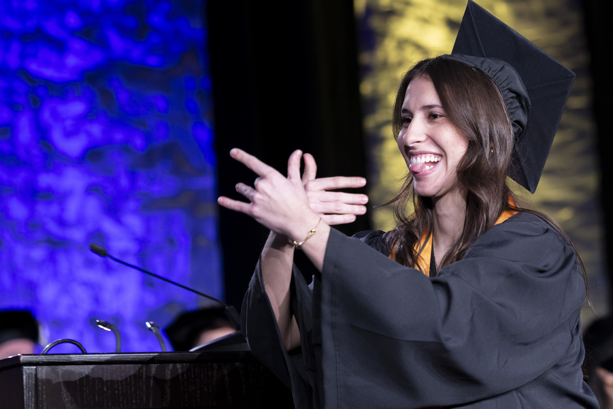 A young woman graduate smiles while gesturing enthusiastically on stage.