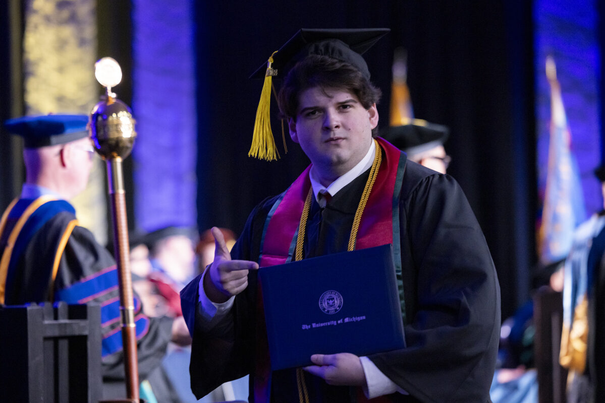 A graduate in cap and gown holds his diploma and points to it proudly.