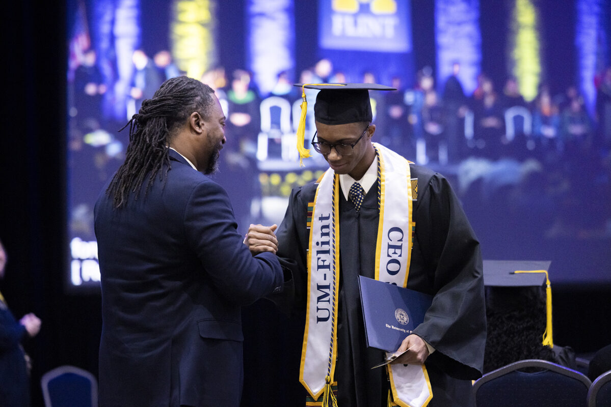 A graduate in a black cap and gown shakes hands with an official on stage.