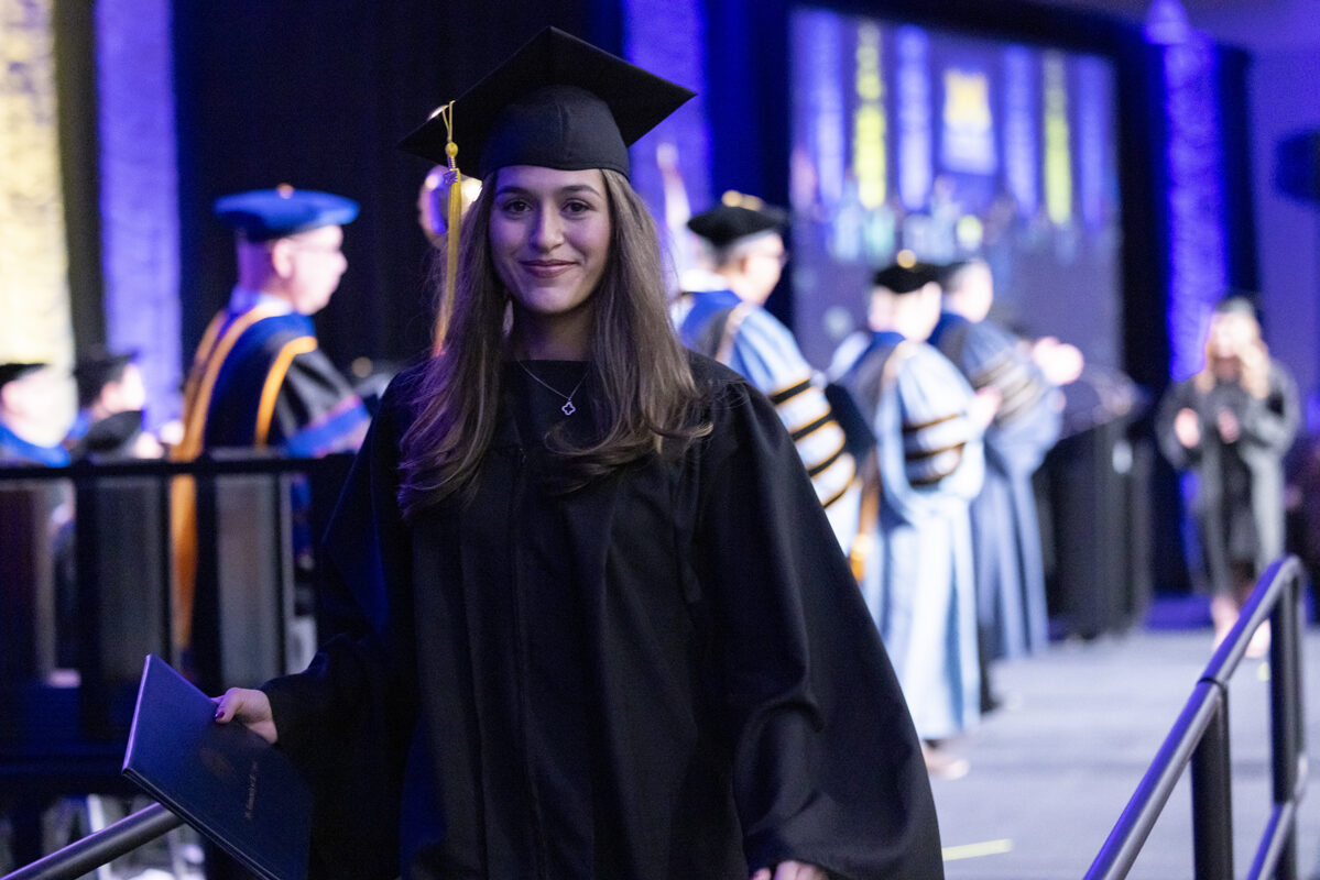 A graduate walks down the stage holding her diploma and smiling.