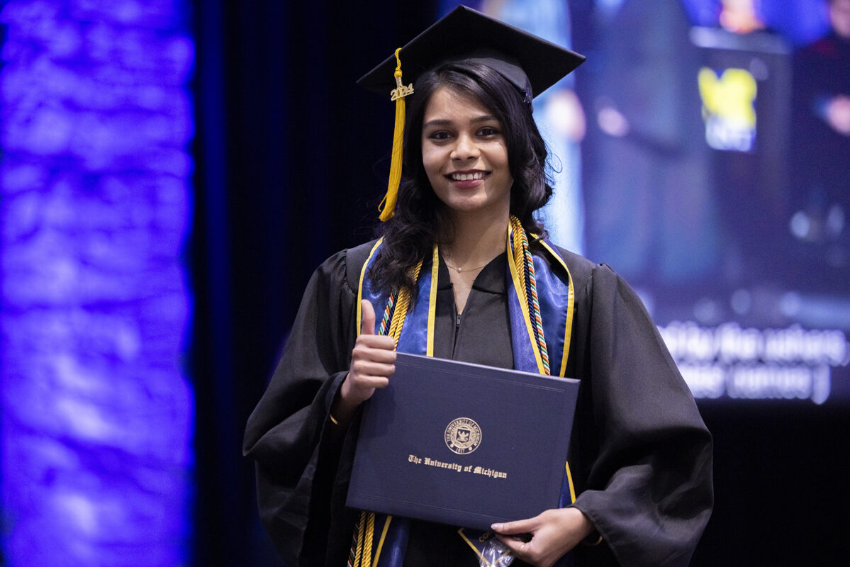 A smiling graduate holds her diploma folder, giving a thumbs-up.