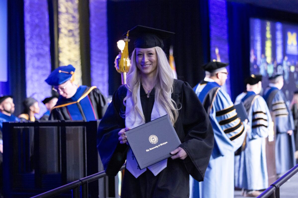 A graduate in cap and gown smiles brightly while holding her diploma.