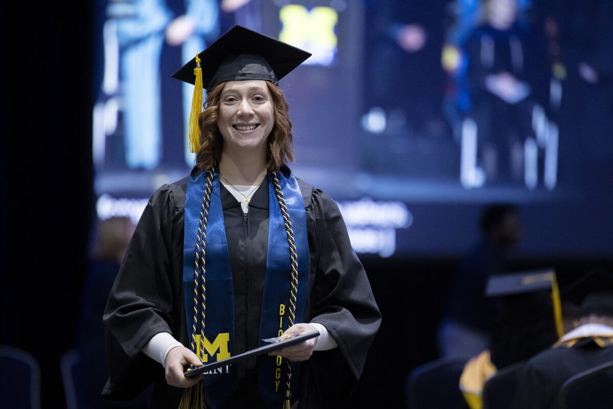 A graduate in a biology stole smiles, holding her diploma.