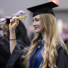 A young woman with long blonde hair in a black cap and gown smiles as she moves the tassel on her graduation cap.