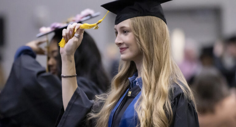 A young woman with long blonde hair in a black cap and gown smiles as she moves the tassel on her graduation cap.