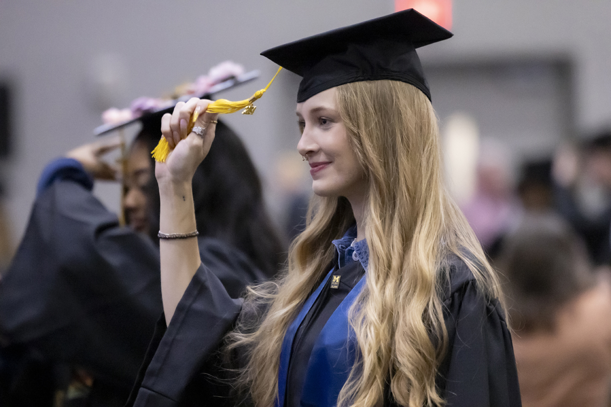 A young woman with long blonde hair in a black cap and gown smiles as she moves the tassel on her graduation cap.