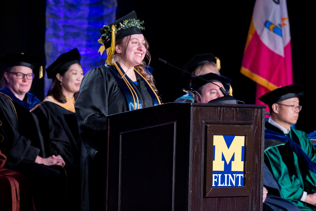 A smiling graduate speaks at a podium with the "M Flint" logo.