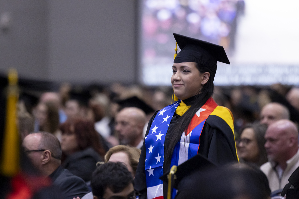 A graduate stands in the audience wearing a red, white, and blue flag-themed stole.