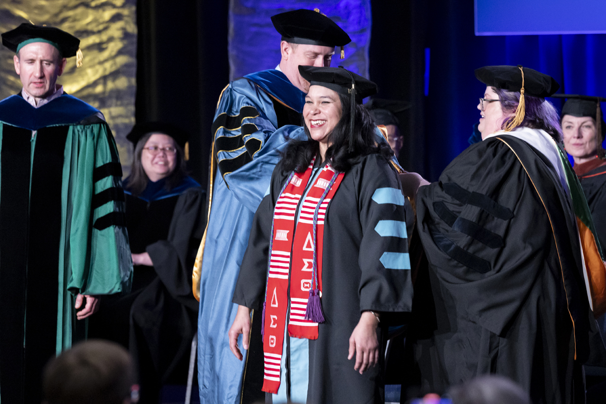 A smiling graduate is hooded on stage during a formal ceremony.