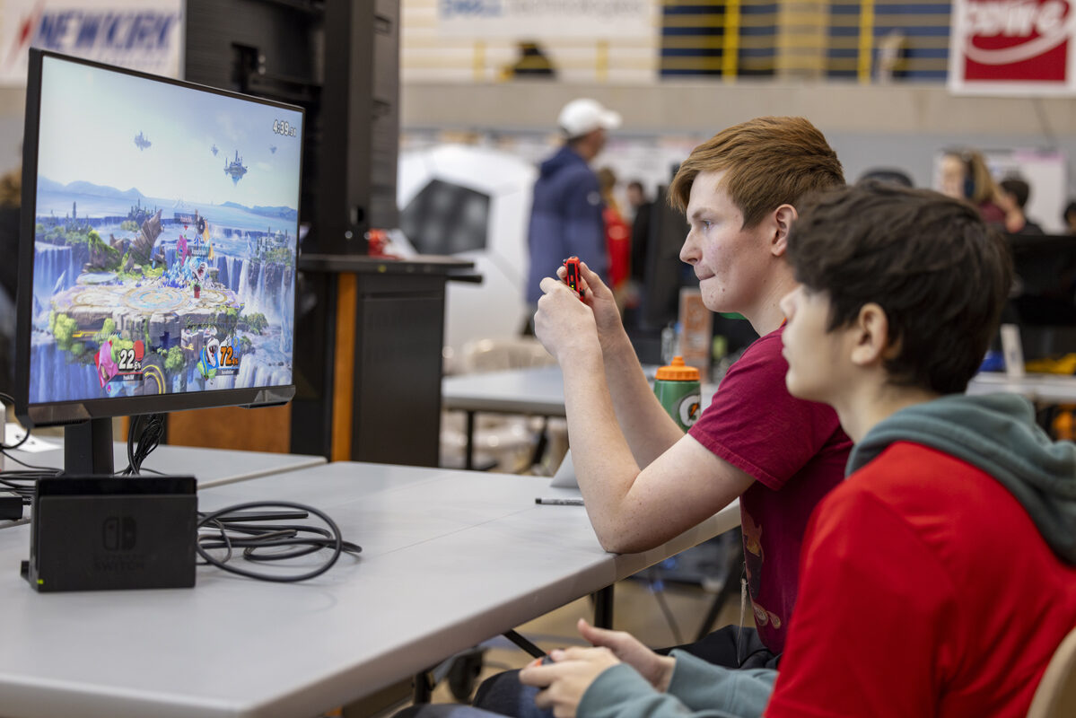 Two people playing a video game on a monitor, seated at a table.