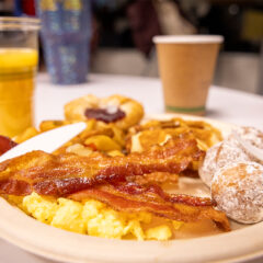 A plate of breakfast food with scrambled eggs, bacon, powdered doughnut holes, and a Danish, accompanied by a cup of coffee and a glass of orange juice.