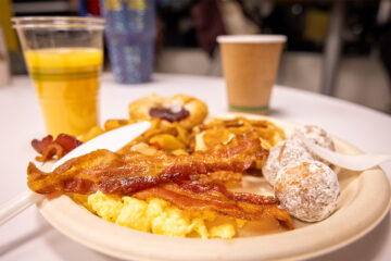 A plate of breakfast food with scrambled eggs, bacon, powdered doughnut holes, and a Danish, accompanied by a cup of coffee and a glass of orange juice.