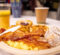 A plate of breakfast food with scrambled eggs, bacon, powdered doughnut holes, and a Danish, accompanied by a cup of coffee and a glass of orange juice.