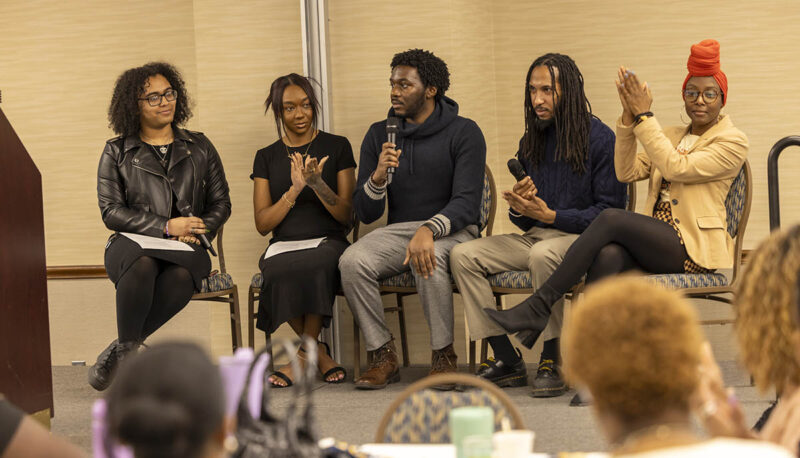 A panel of UM-Flint students seated on stage speaking into a microphne